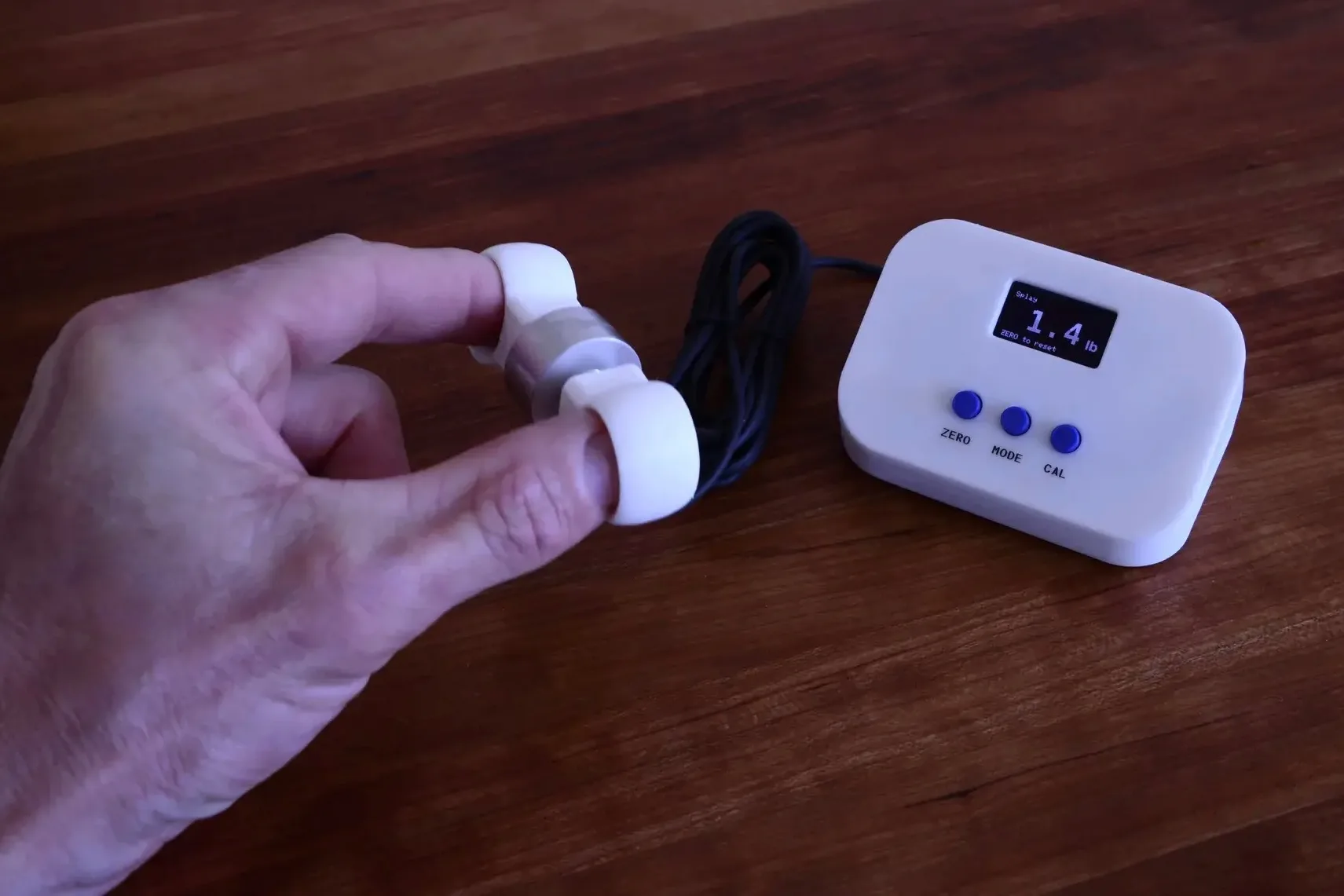 Hand using the Splayometer finger rings, device displaying a real-time force reading of 1.4 lb